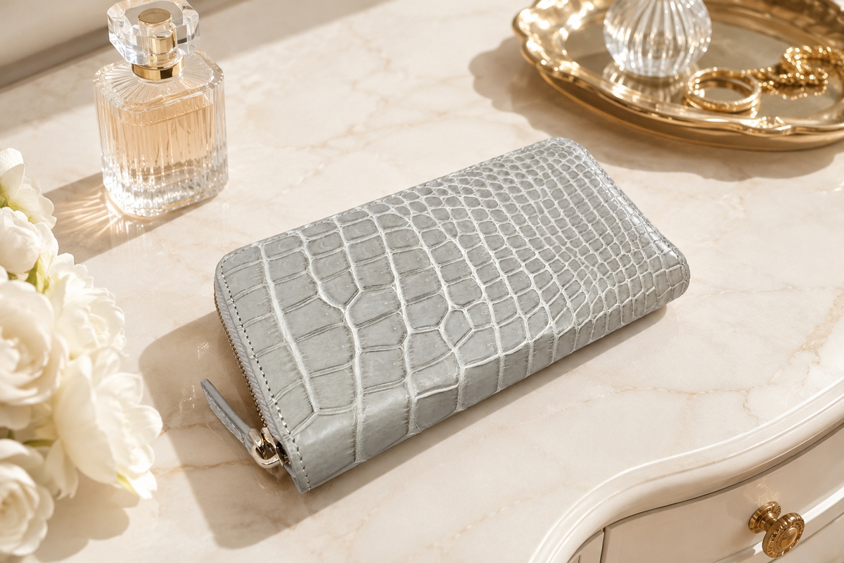 Belvedere Siamese crocodile zip-around wallet in Stone Gray styled on a luxury vanity with perfume, white flowers, and gold accessories