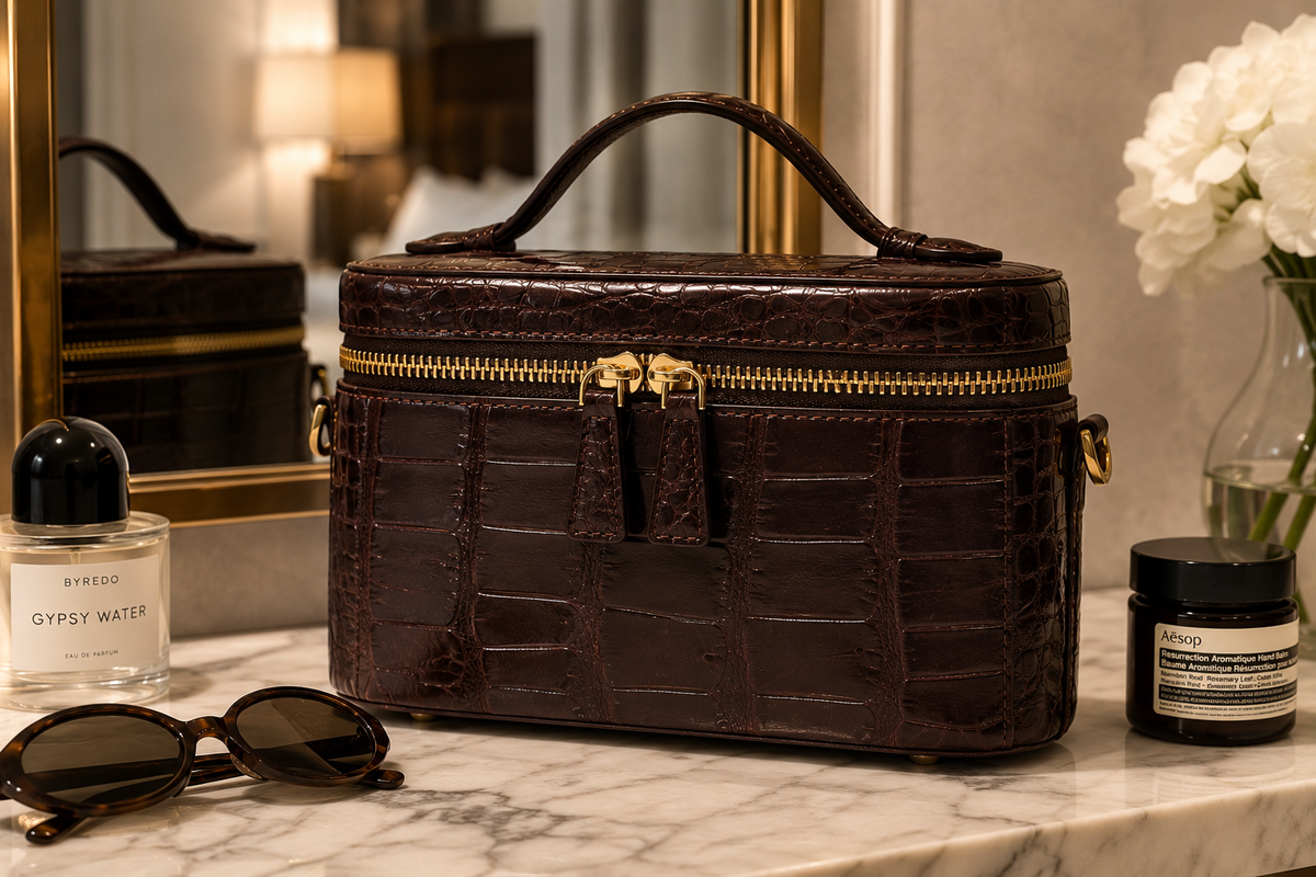 Bordeaux Clarendon crocodile mini box bag photographed on a luxury hotel vanity with perfume and sunglasses, showcasing a structured travel case design and polished gold-tone hardware.
