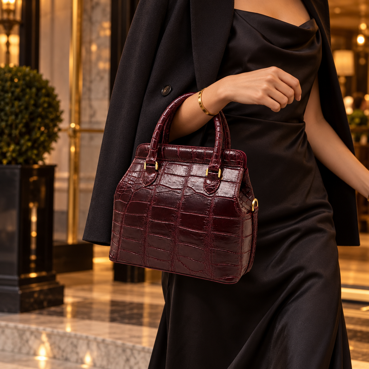 Woman carrying The Ellery Crocodile Handbag in deep Bordeaux by its rolled handles, styled with an elegant black outfit at a luxury hotel.