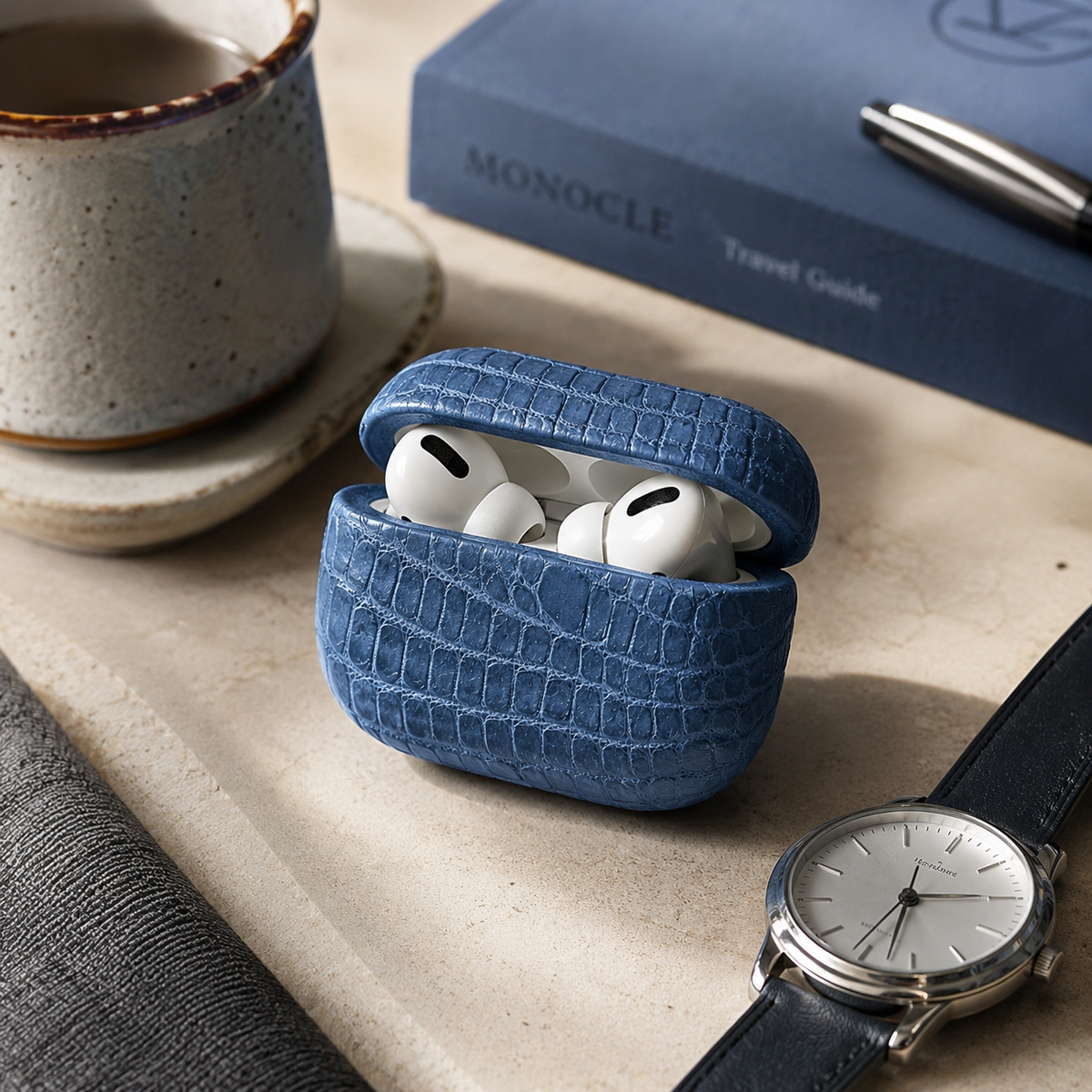 The Crocodile AirPods Case in French Blue genuine Siamese crocodile leather displayed in a luxury desk setting with AirPods, watch, coffee cup, and travel accessories.