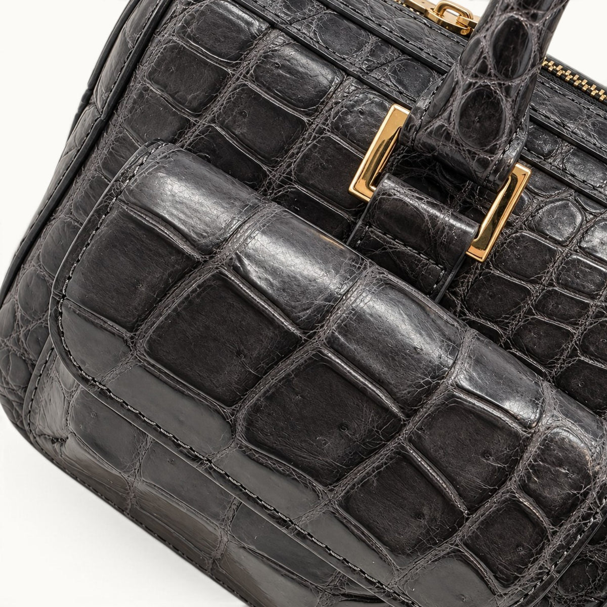 Close-up detail of The Fairmont Crocodile Handbag in gunmetal highlighting the crocodile leather scale texture, front pocket craftsmanship, stitching, and polished gold-tone handle hardware.