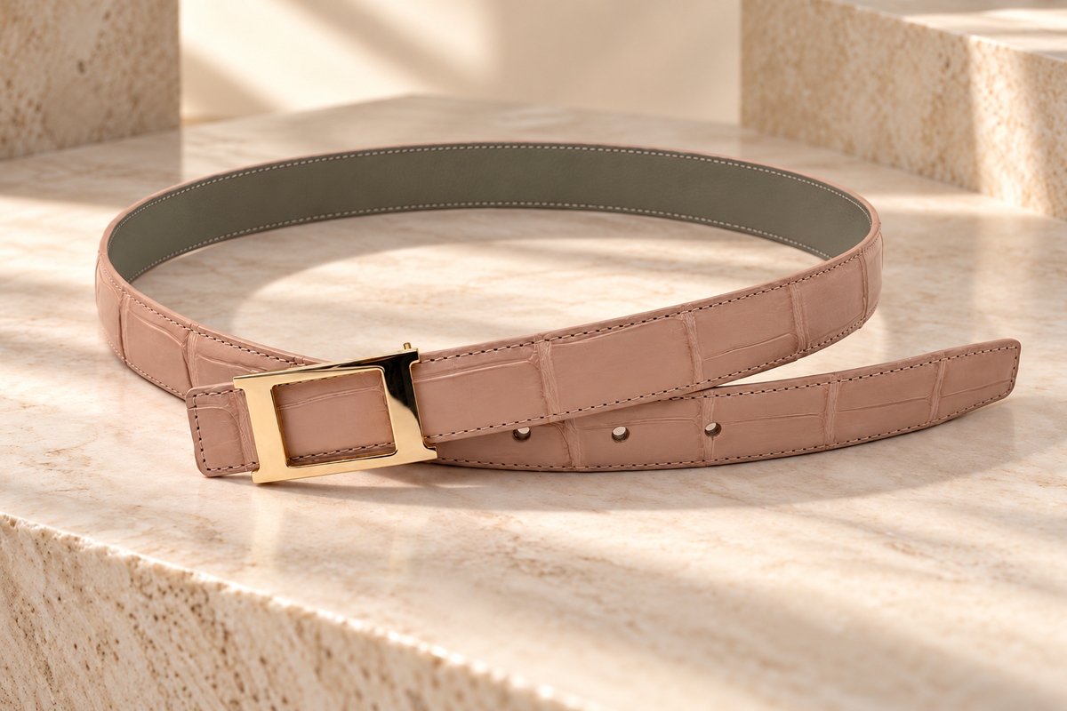 Blush pink crocodile leather belt displayed on natural stone, featuring detailed stitching, olive-gray leather backing, and a gold-tone buckle.
