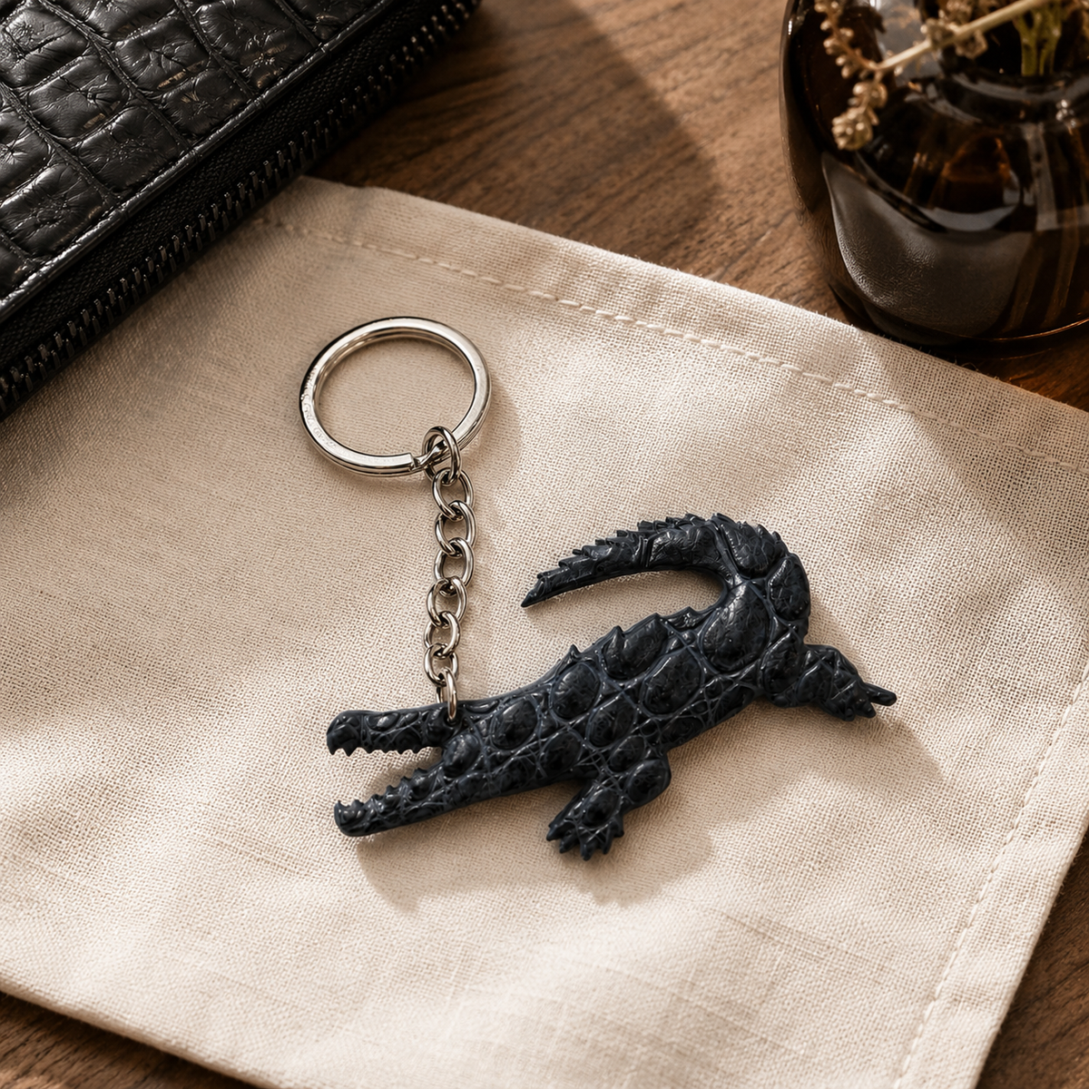 Gunmetal gray Siamese crocodile leather key charm styled on natural linen beside a black leather accessory