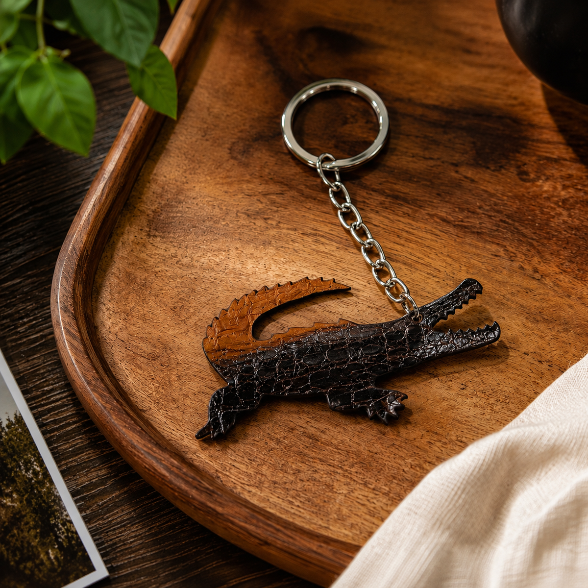 Two-tone brown Siamese crocodile leather key charm styled on a wooden tray with natural linen