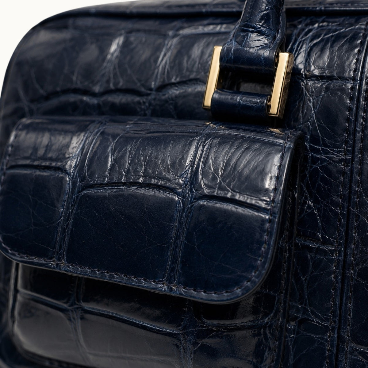 Close-up detail of The Fairmont Crocodile Handbag in navy blue highlighting crocodile texture, front pocket, stitching, and gold-tone handle hardware.