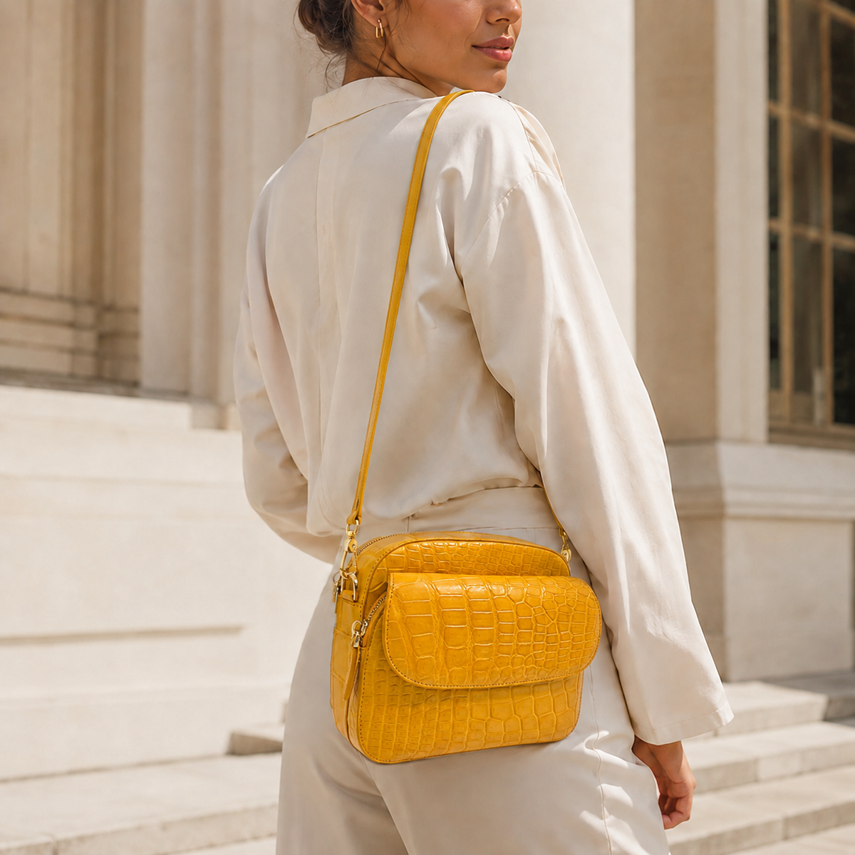 Woman wearing the Grosvenor Siamese crocodile crossbody bag with an ivory outfit outside an elegant stone building.