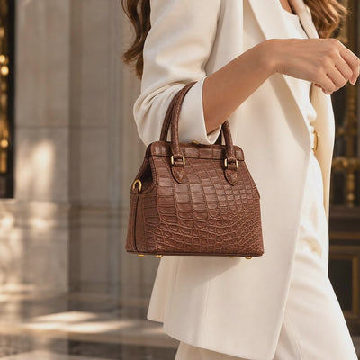 Woman carrying The Ellery Crocodile Handbag in Cocoa Brown with an elegant ivory outfit, showcasing its natural crocodile leather texture, rolled handles, and gold-tone hardware.