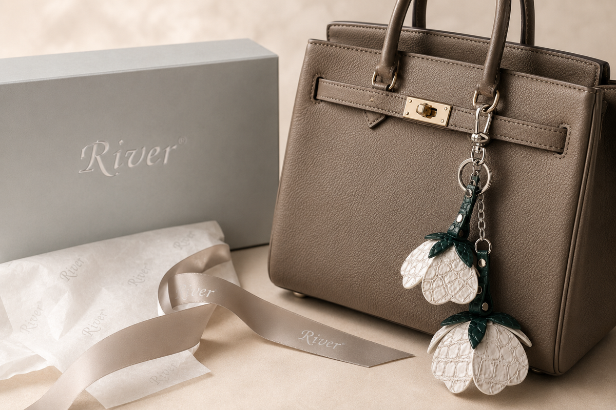 Stone Ivory Valencia crocodile flower charm hanging from a taupe handbag beside River luxury gift packaging